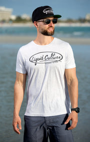 A man standing on a beach wearing a white t-shirt with a black 'Liquid Culture' logo, a black cap, and sunglasses.