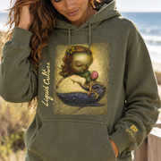 Woman wearing an army green Liquid Girl hoodie featuring whimsical girl and whale artwork on a beach.